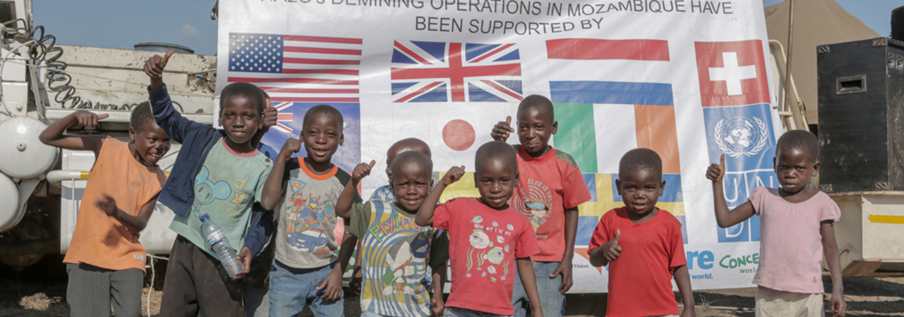 Children stand and smile in front of a banner celebrating a Mine Free Mozambique, with flags of donor countries including US, UK, Ireland, and Japan