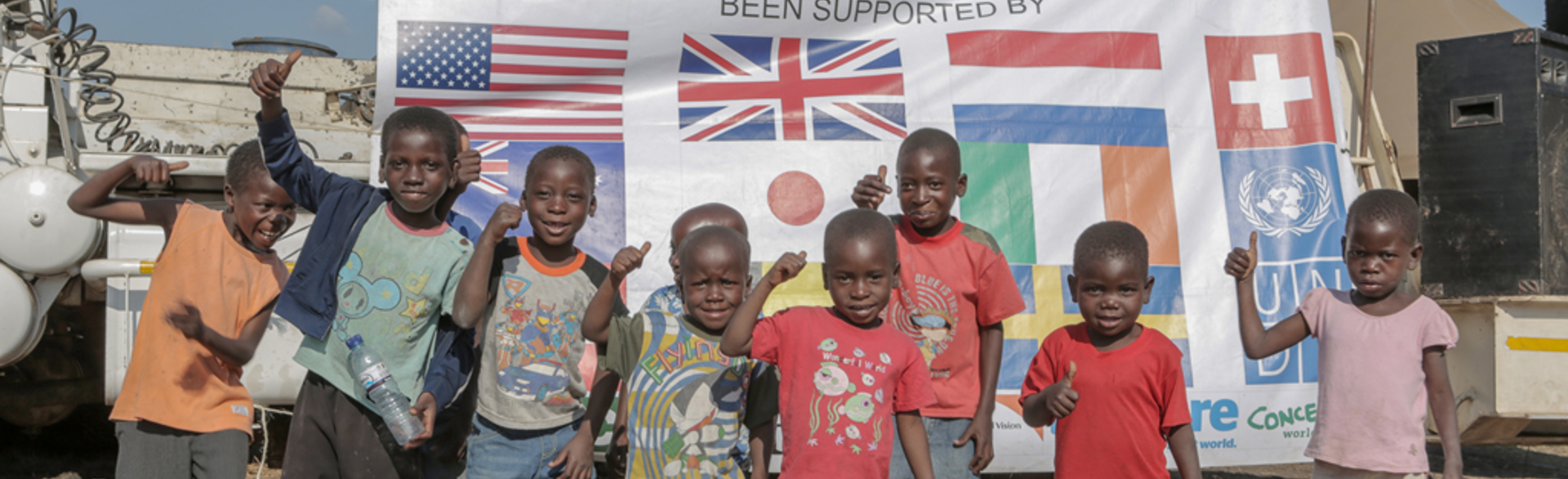 Children stand and smile in front of a banner celebrating a Mine Free Mozambique, with flags of donor countries including US, UK, Ireland, and Japan