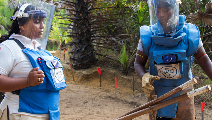 Two deminers hold vegetation cutting tools in Sri Lanka whilst wearing personal protective equipment and visors