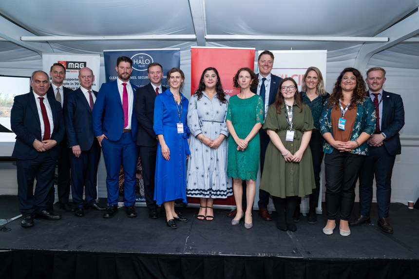 A group of MPs stand on the stage in front of the MAG, HALO, and Labour Party Banners at the Labour Party Conference