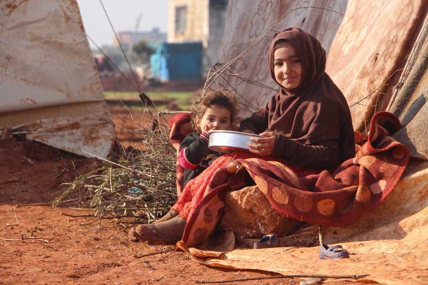 A child and a toddler sit on the ground holding a metal bowl