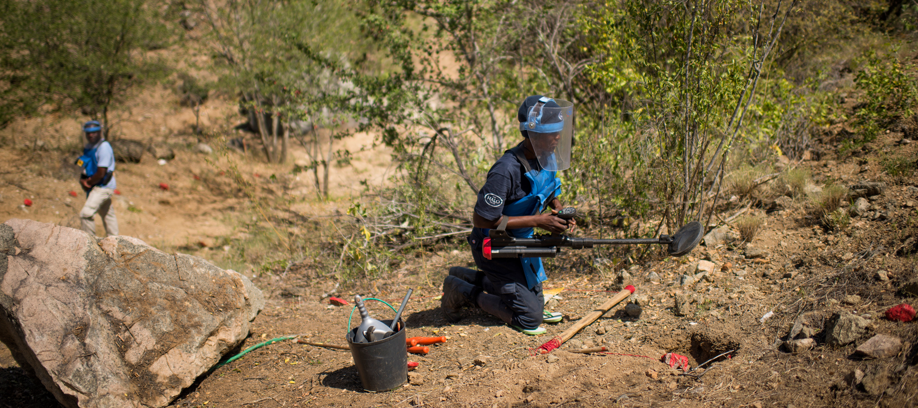 A female deminer kneels down on the soil whist holding a metal detector