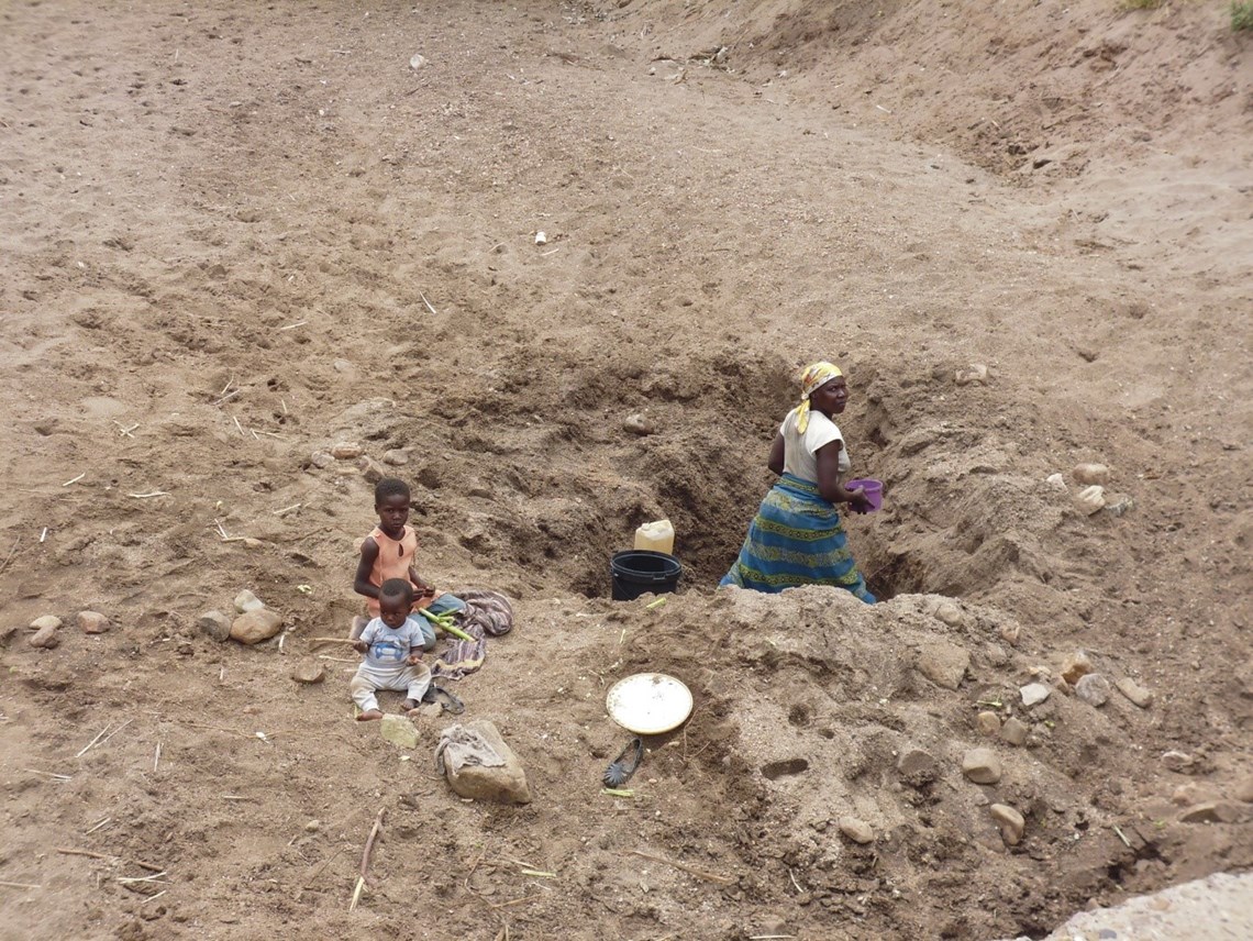 A mother digs for water in a dry river bed near to HALO’s second camp as her young child and baby look on, HALO Trust.