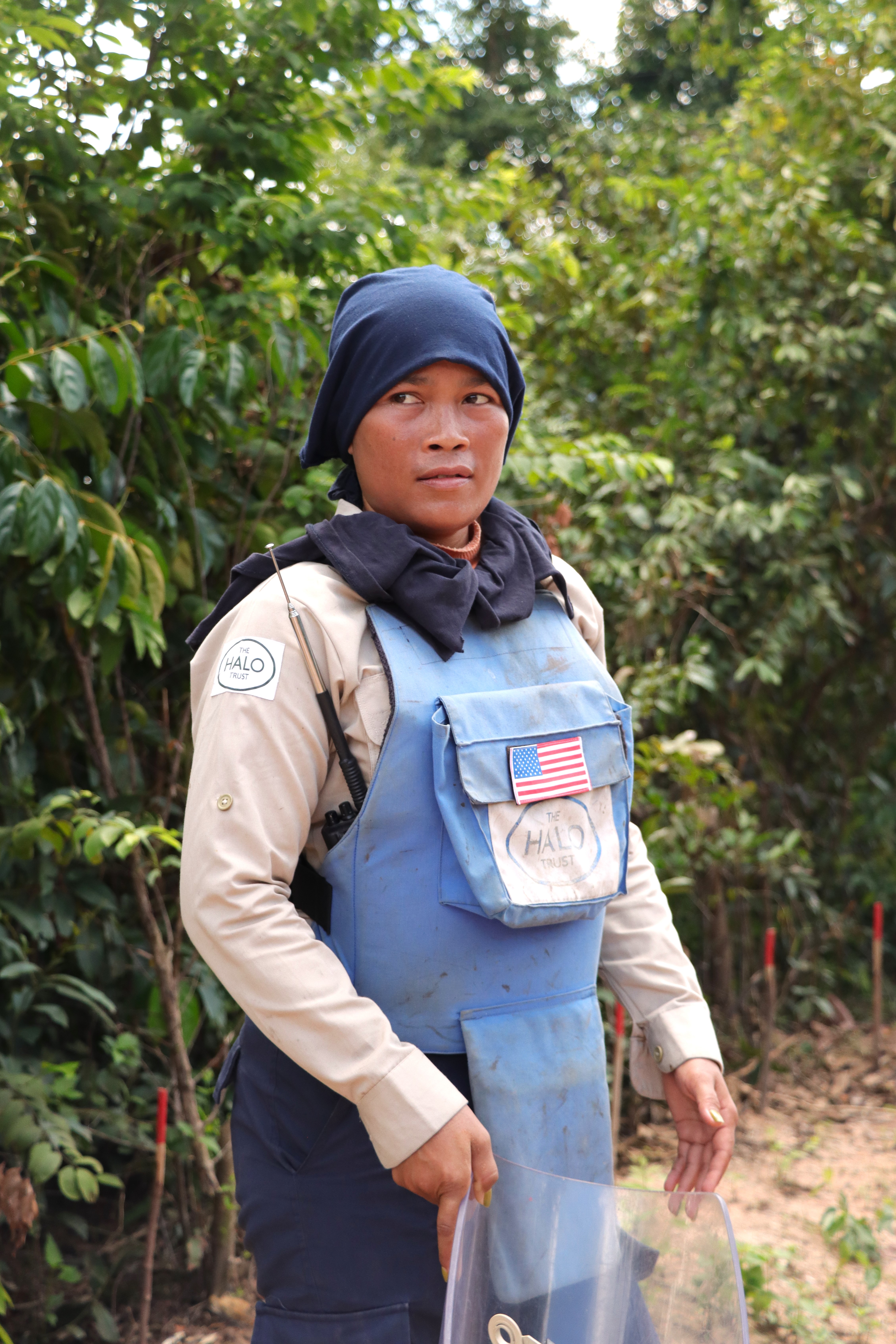 A female deminer stands in a blue vest with an American flag against a green background.