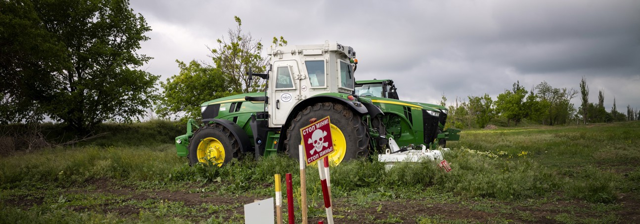 A large agricultural machine stands in the middle of a field behind a landmine warning sign.