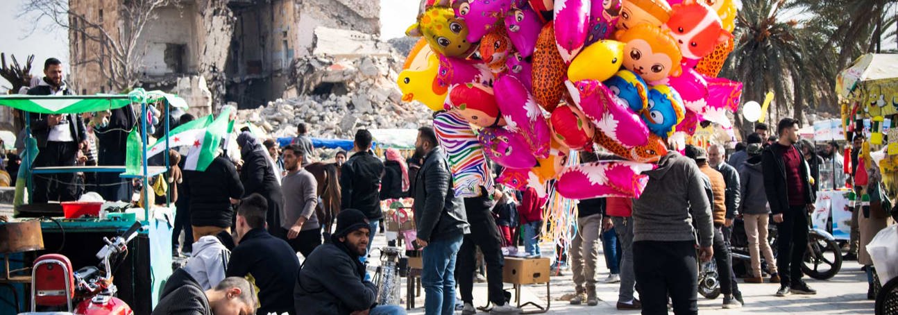 A destroyed building stands in the distance as crowds walk through the Alepp Citadel