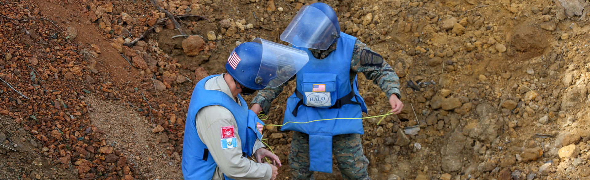 Deminers wearing PPE hold wires as they carry out a bulk demolition on an unexploded ordnance