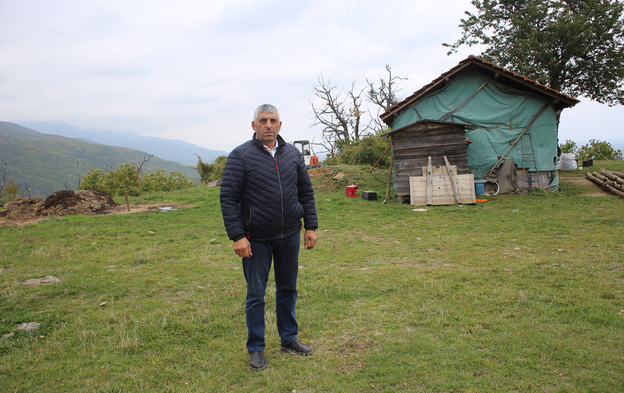A man stands in a green yard with a small house in the background and mountains in the distance.