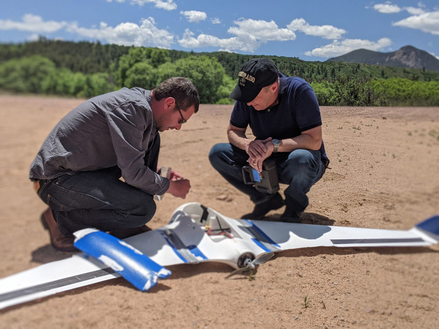 Two men crouch on the ground while examining a revolutionary drone used to detect landmines and explosive ordnance