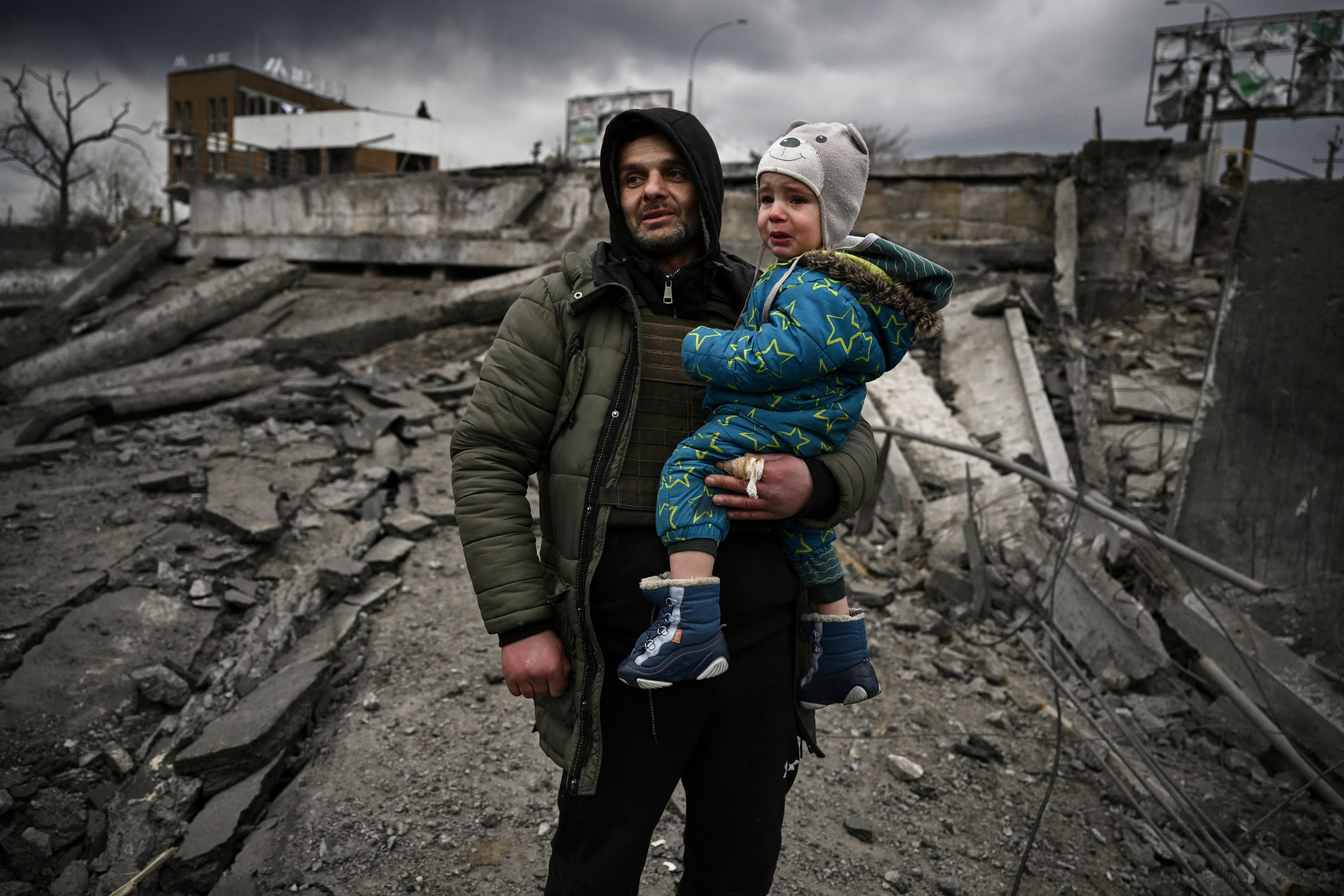A man holds a crying child in his arms with destroyed buildings behind him