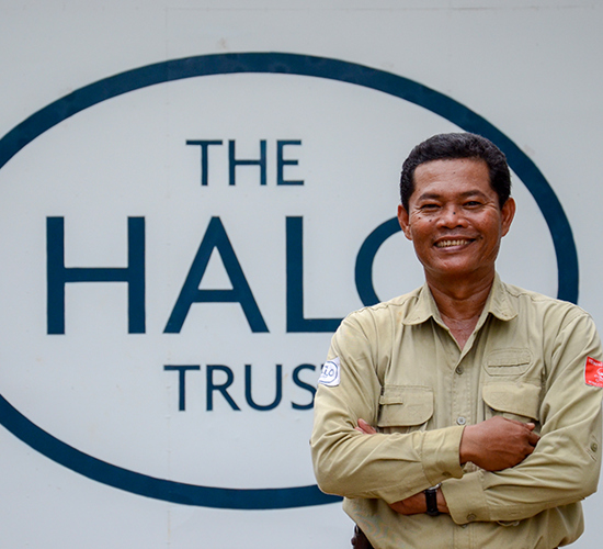 Mr Smann Makara, stands in a HALO shirt in front of The HALO Trust logo