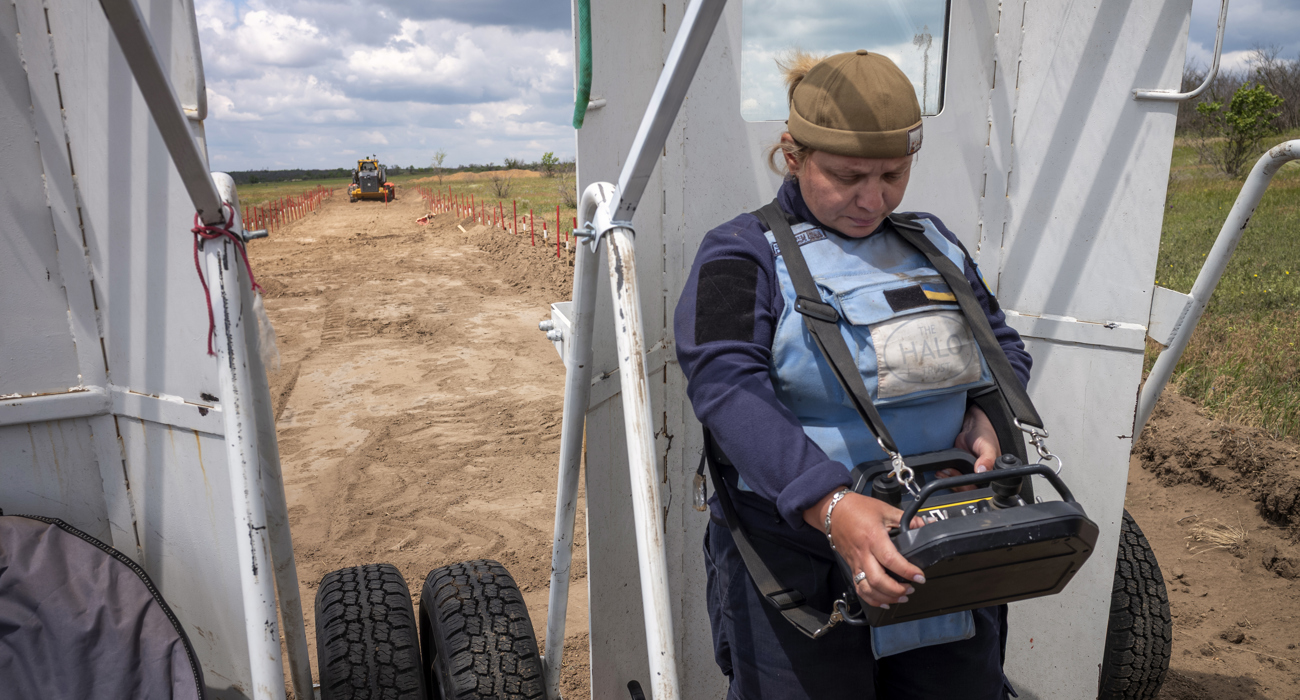 A woman in a HALO Trust vest operates a large remote control, controlling a large vehicle from a distance. 