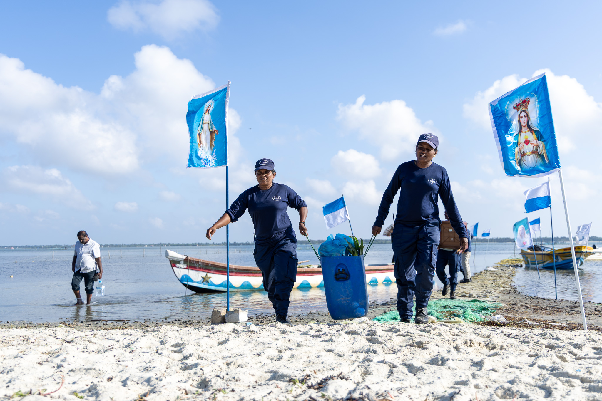 Two women hold a bag of supplies on each side, their backdrop is a blue lagoon and two floating boats.