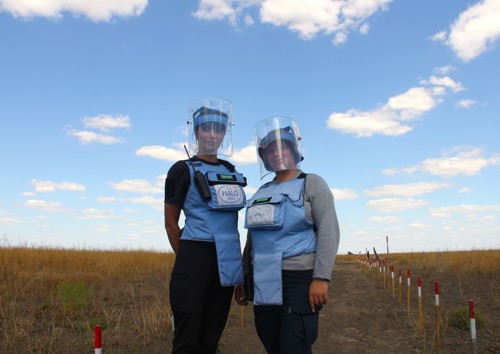 Two female deminers pose for a photo in a field with a blue sky behind them.