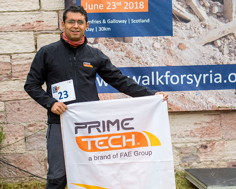 Emilio Canales from Primetech, holds a Primetech banner while standing in front of a walk for Syria banner