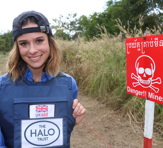 Camilla Thurlow stands in PPE in front of a Danger Mines sign in a field in Cambodia during her training