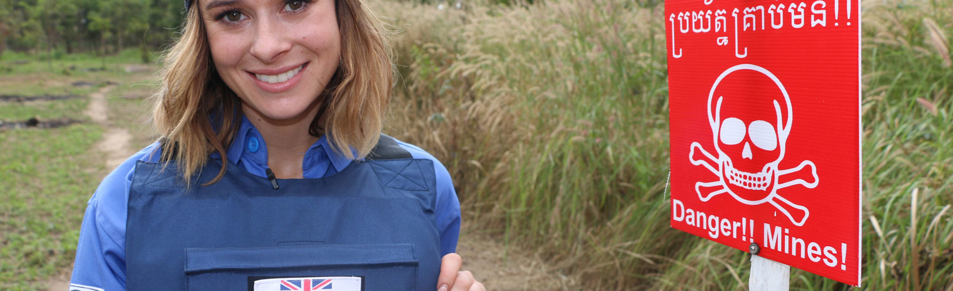 Camilla Thurlow stands in PPE in front of a Danger Mines sign in a field in Cambodia during her training