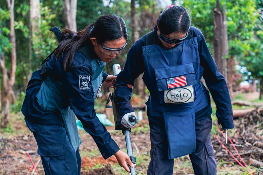 Two female deminers in PPE use a detector in a forest