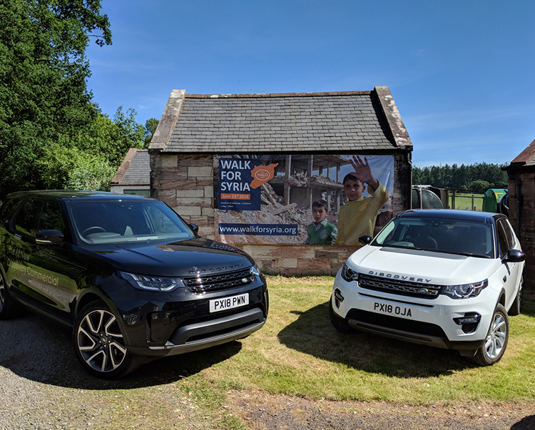 Two shiny new Landrover Discoveries outside a house with a walk for Syria banner