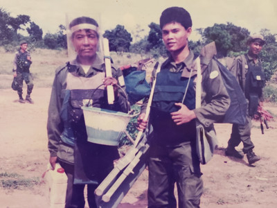 A photo of two deminers in Cambodia wearing PPE and carrying equipment in a field in Cambodia
