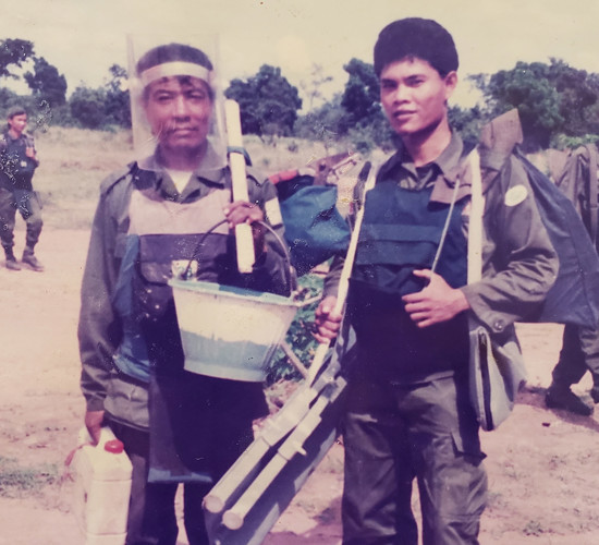 Two deminers in Cambodia wearing PPE and carrying equipment in a field in Cambodia
