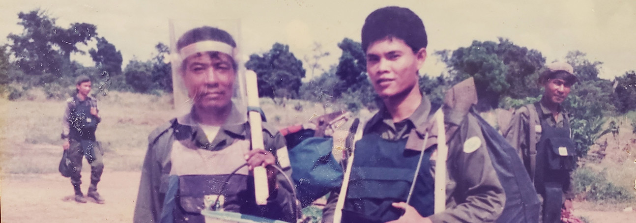 Two deminers in Cambodia wearing PPE and carrying equipment in a field in Cambodia