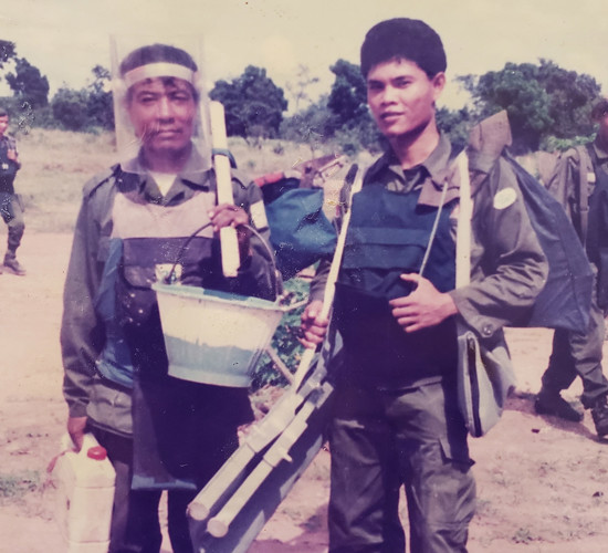 A photo of two deminers in Cambodia wearing PPE and carrying equipment in a field in Cambodia