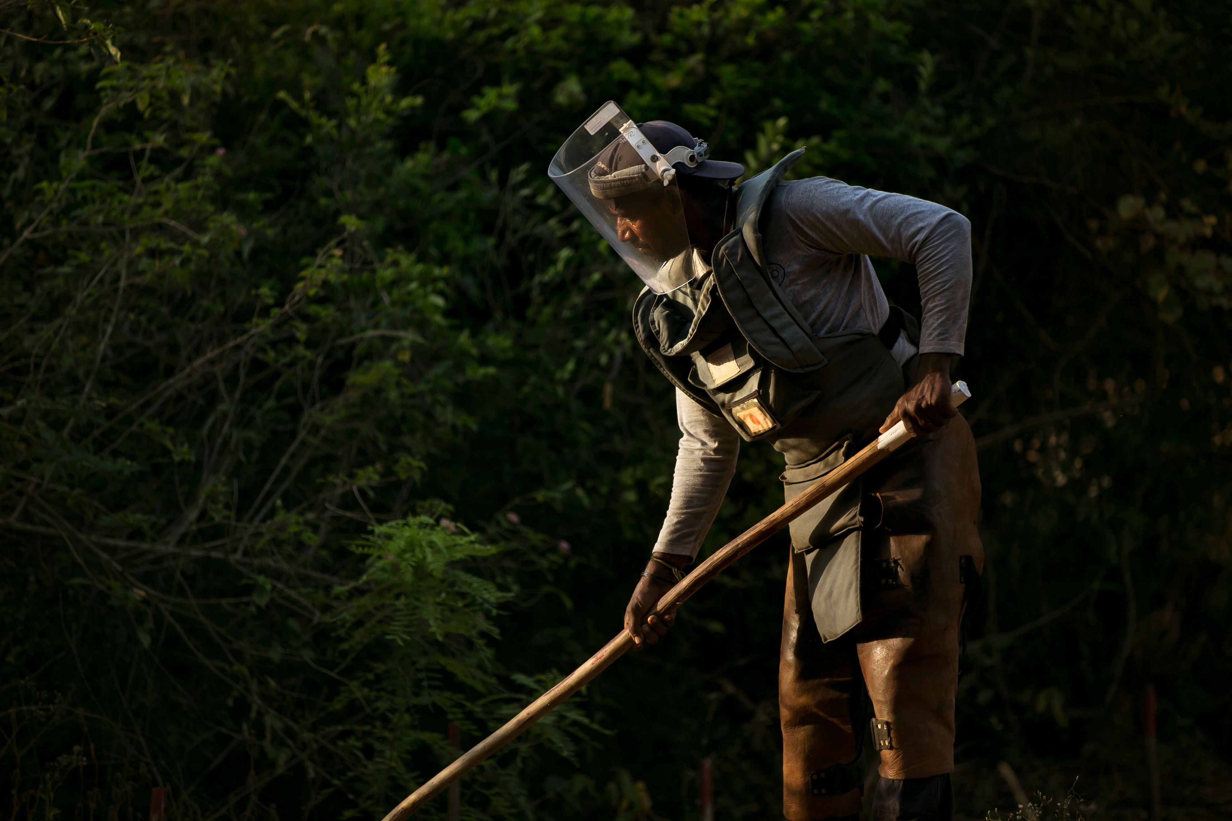 A woman in full demining PPE works standing in front of lush green vegetation