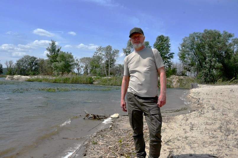 A man in a gray shirt and a cap stands on the shore of a lake.