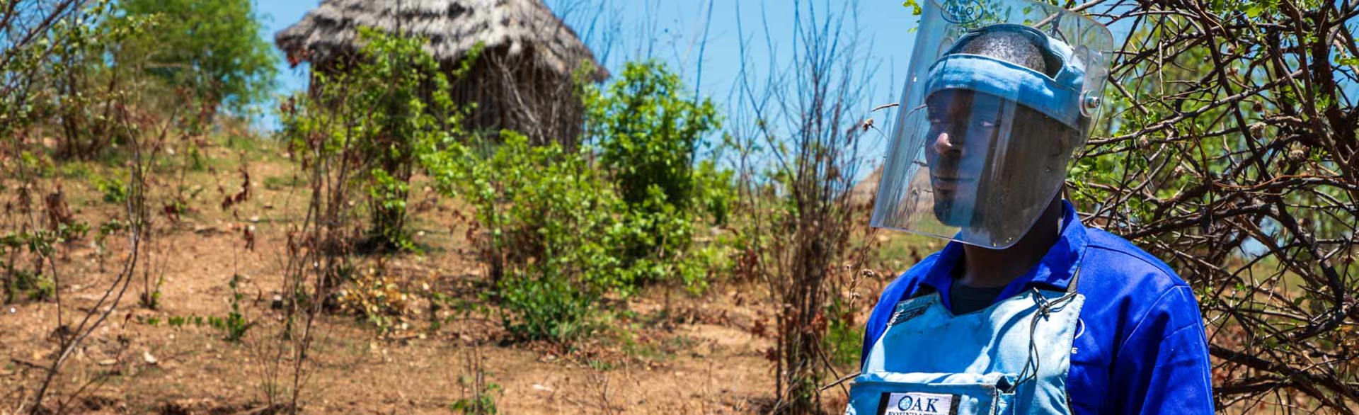 A HALO deminer wearing PPE and a visor next to a village in Zimbabwe