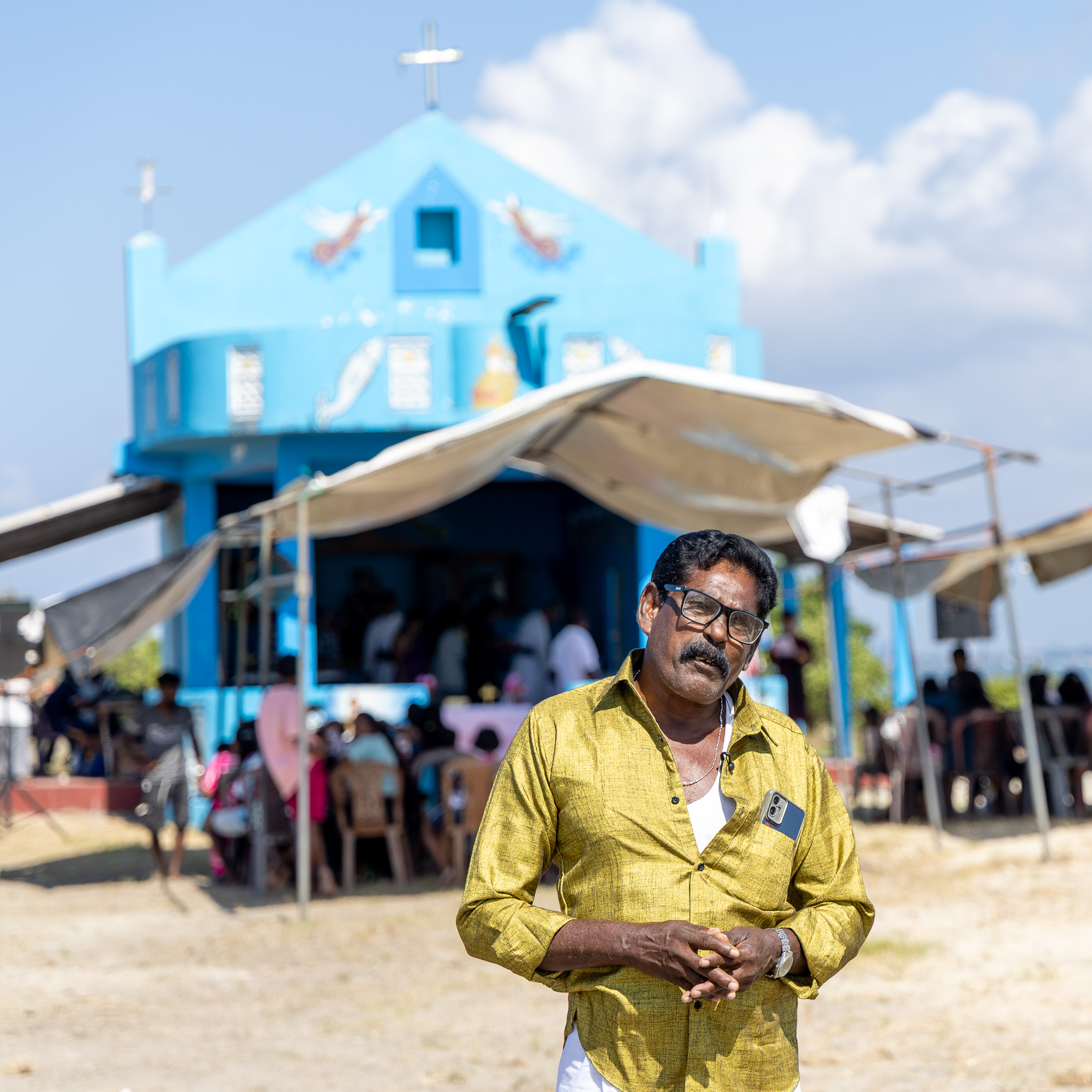 A man dressed in a yellow shirt stands with his hands knitted together in front of a blue church.