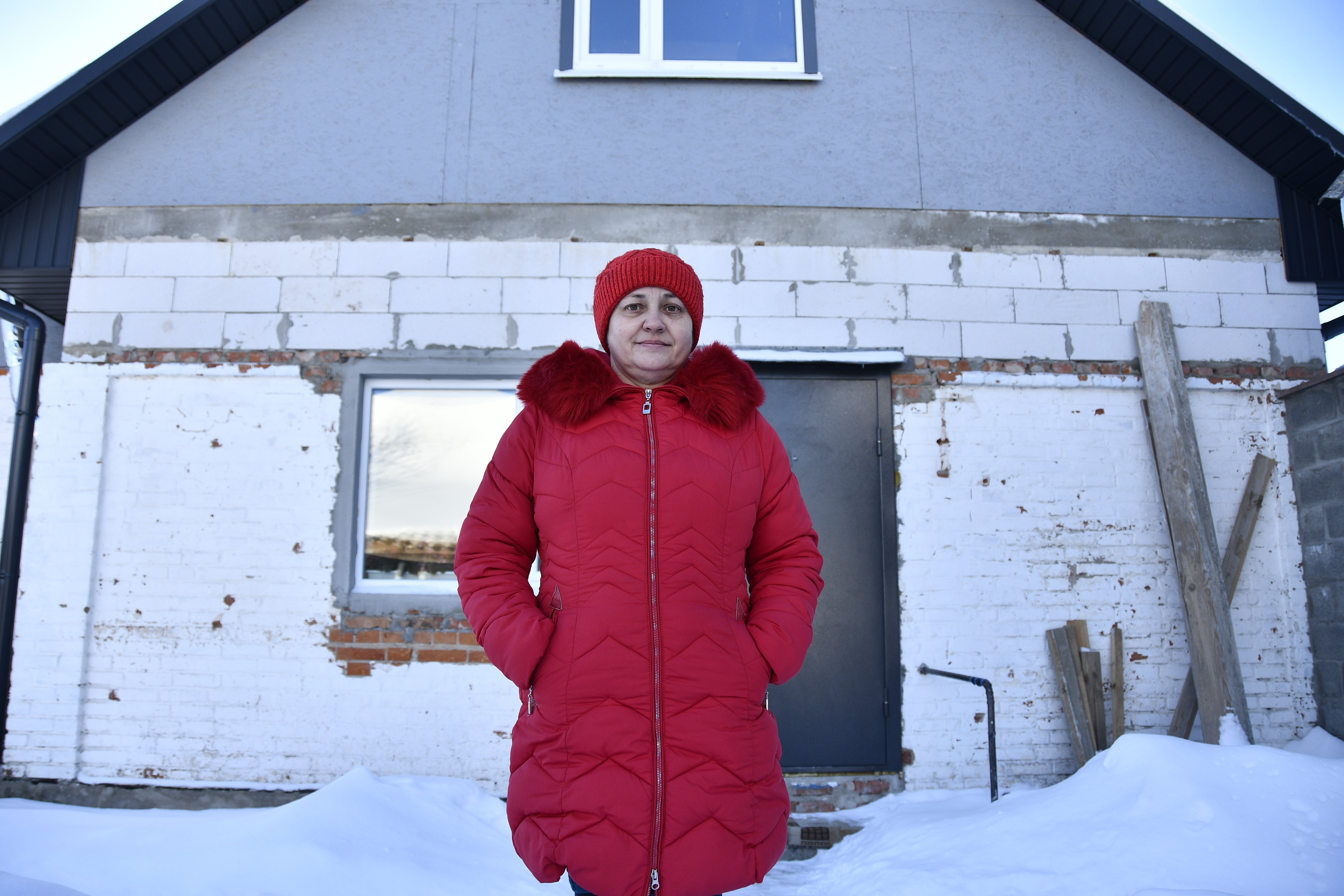 Oksana stands in her red hat and coat outside her white house.