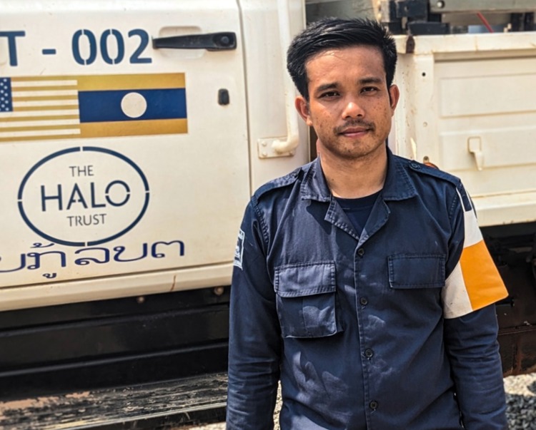 Nathana Kedtavong, mine clearance team leader, stands in uniform in front of a HALO vehicle in Laos