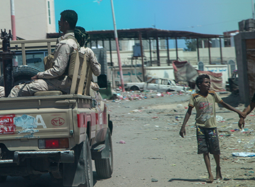 Military personnel ride in the back of a pick truck while a young boy walks through the littered streets of Yemen