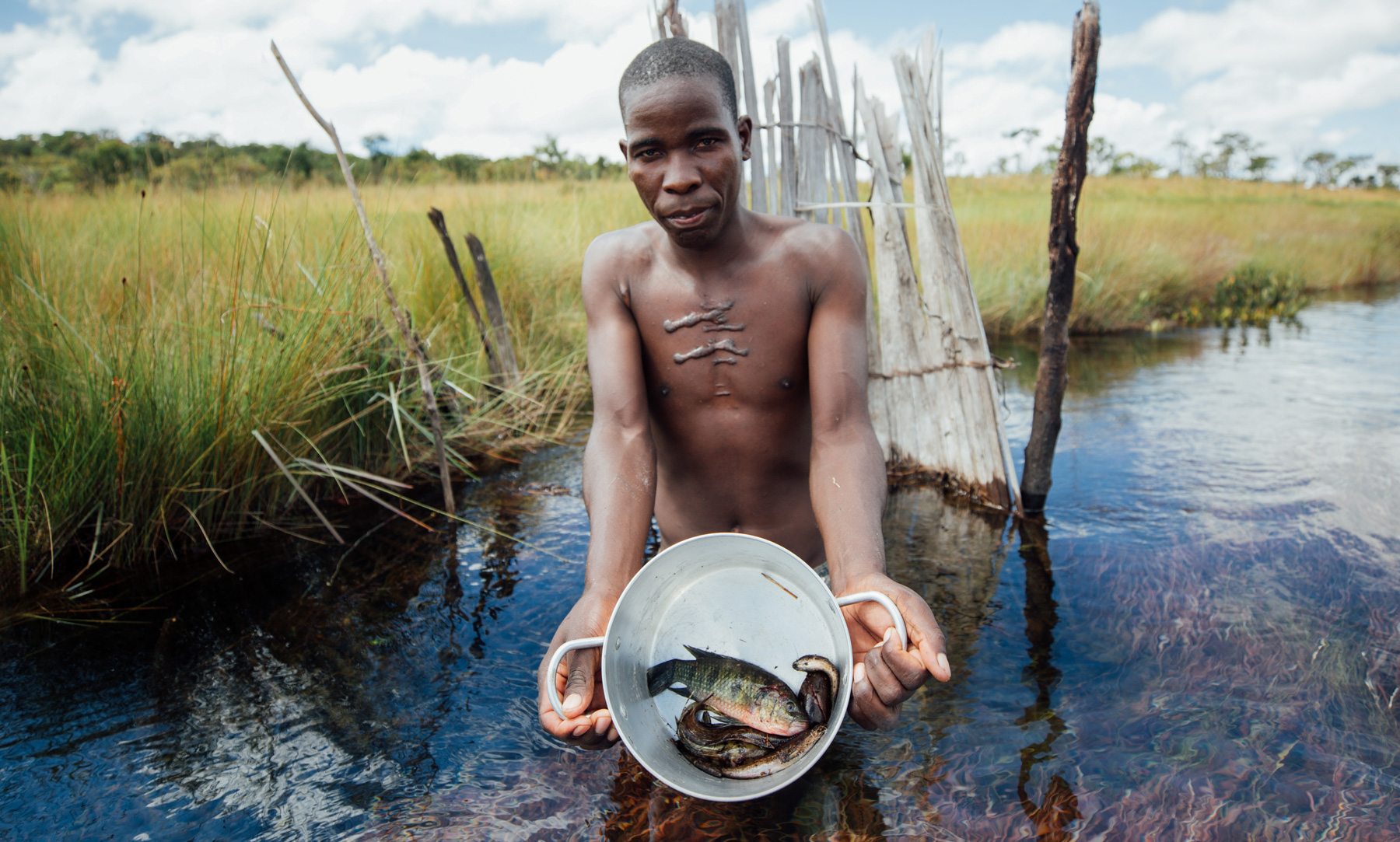 A fisherman stands in the river and shows a bucket of fish