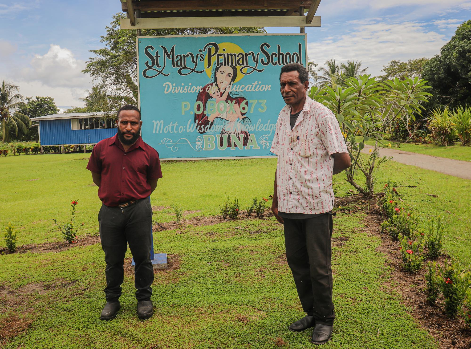 Two male teachers stand outside in front of a sign that reads St. Mary's Primary School, Buna.