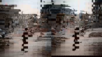 Woman dressed in black, walking through a street in Syria surrounded by rubble from destroyed buildings