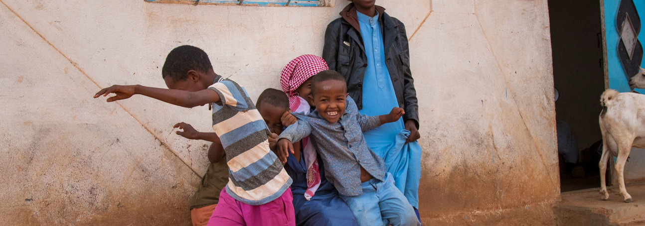 A group of children play outside a building next to a smiling adult and a goat in Somaliland 