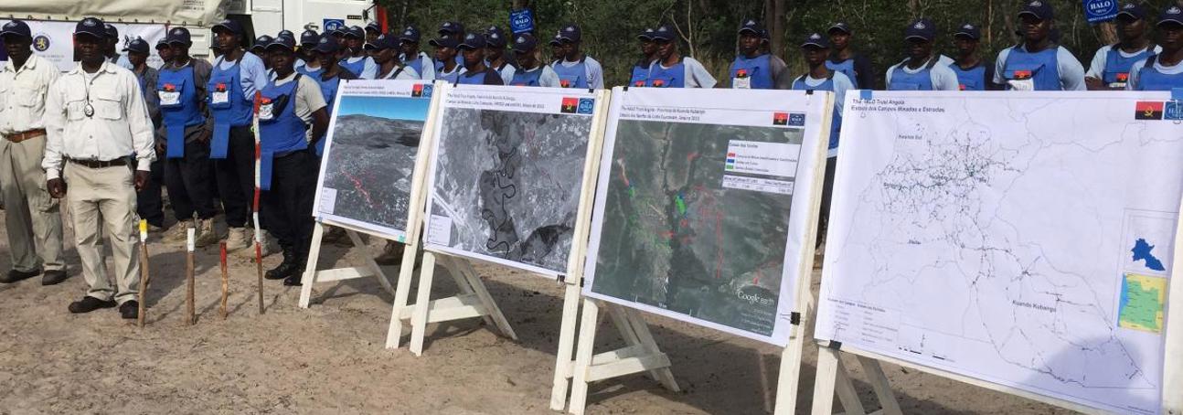 A large group of HALO Trust deminers stand behind boards displaying images and maps highlighting the contmination following the battle of the Cuito Cuanavale