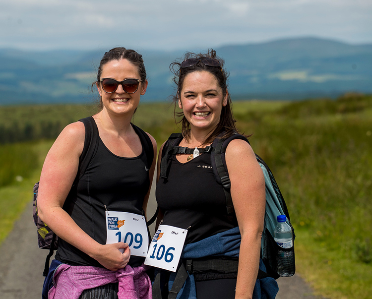 Two women smile with their badge numbers as they walk for Syria