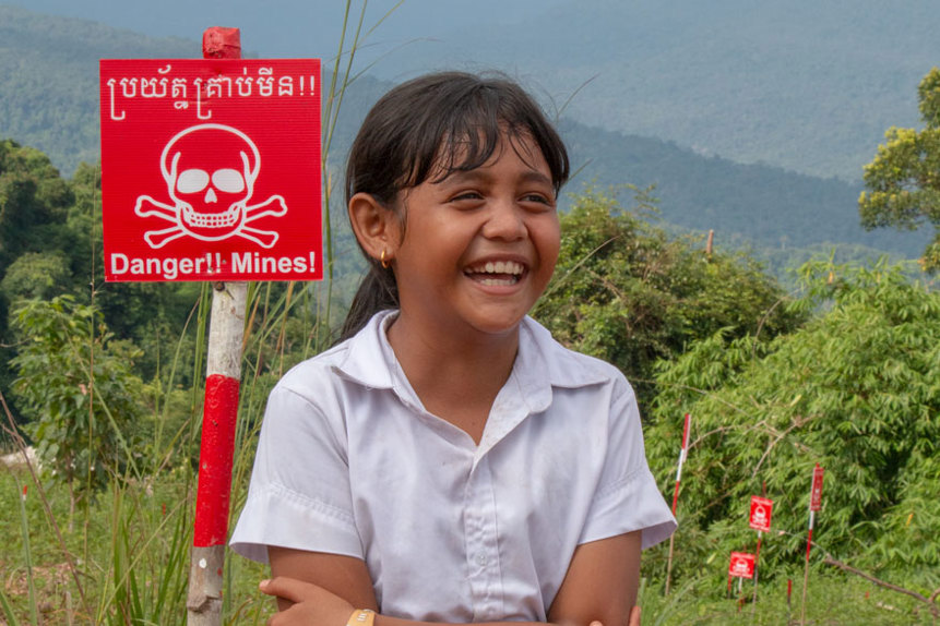 A girl sits in front of a danger mines sign in a field in Cambodia