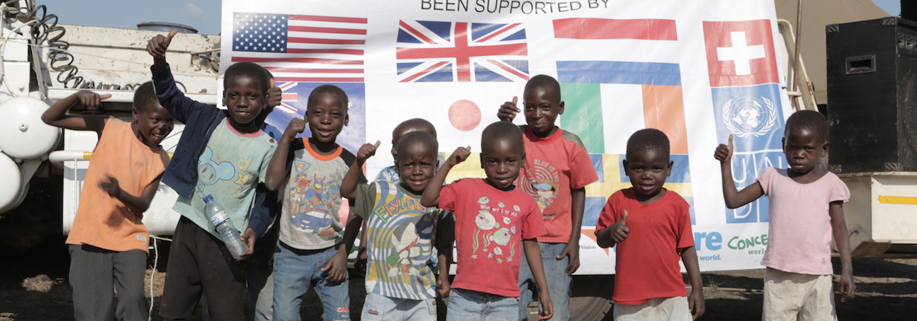A large group of children smile and give a thumbs-up to the camera in front of a banner declaring Mozambique's mine free status