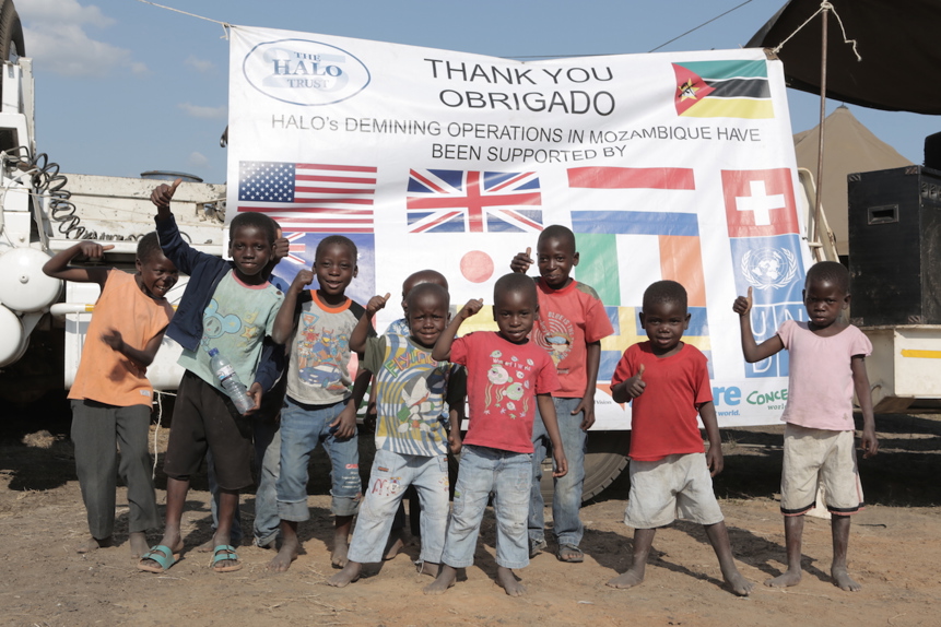 A large group of children smile and give a thumbs-up to the camera in front of a banner declaring Mozambique's mine free status