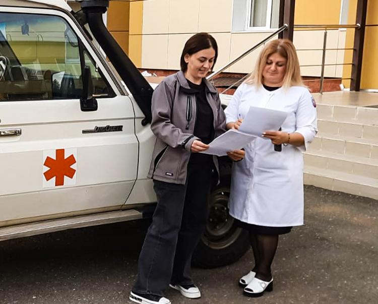 Two women stand in front of a van holding papers
