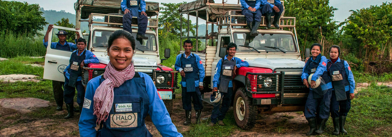 A group of deminers in Cambodia smile in front of their vehicles wearing PPE