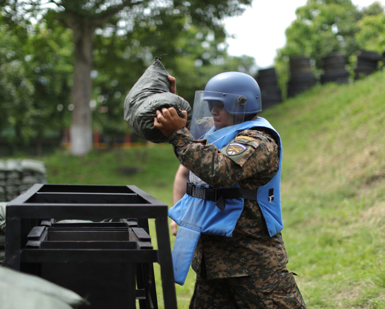 A HALO Trust Weapons and Ammunition technician holds a large sandbag whilst wearing a protective helmet and visor