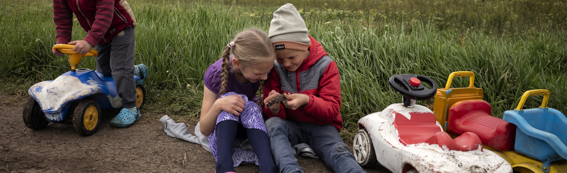 Two young children play together in a field. One boy rides a toy car to the left side.