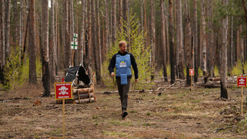 Prince Harry, wearing a HALO vest with the Ukrainian flag, walks through a wooded area featuring a "danger: mines" sign.