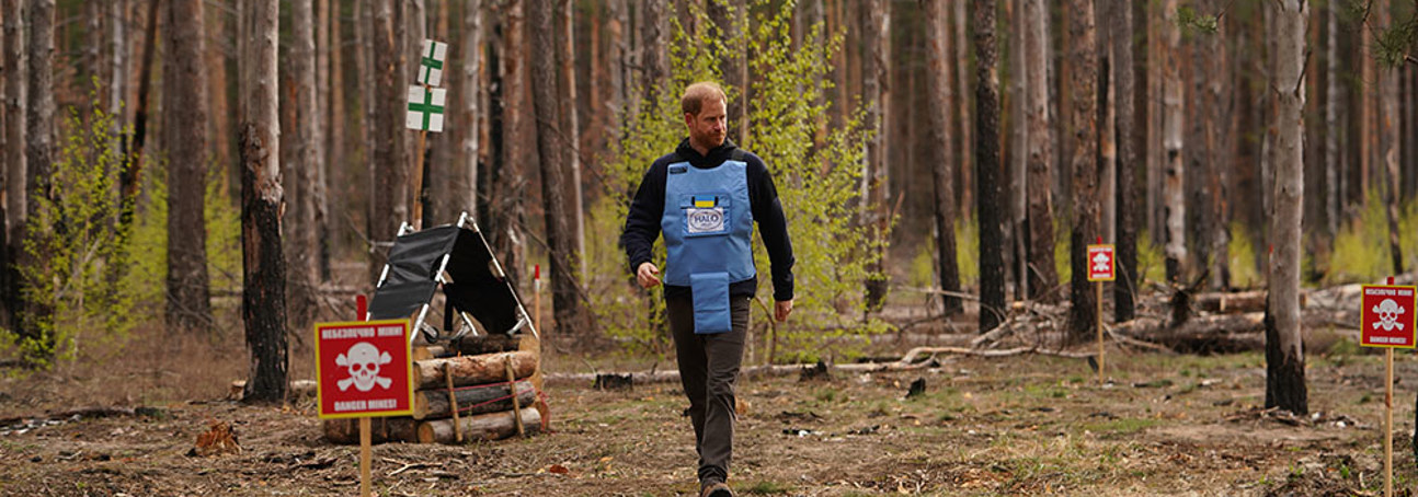 Prince Harry, wearing a HALO vest with the Ukrainian flag, walks through a wooded area featuring a "danger: mines" sign.