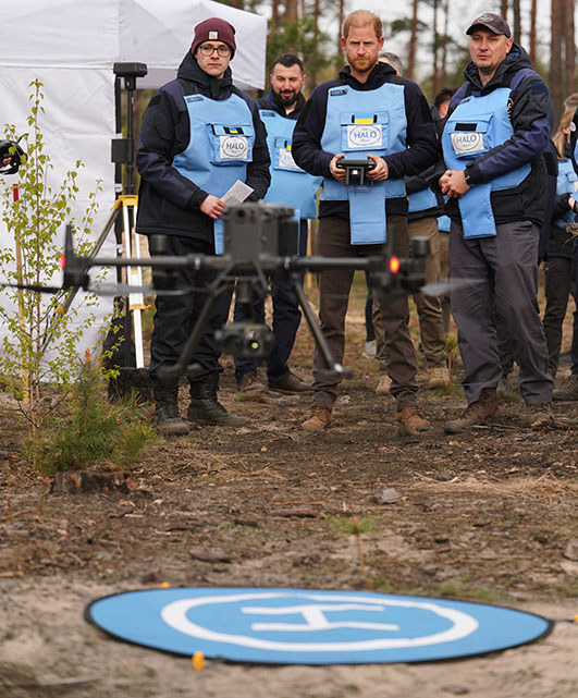 Prince Harry, surrounded by HALO Ukraine team members, pilots a drone which hovers off the ground.